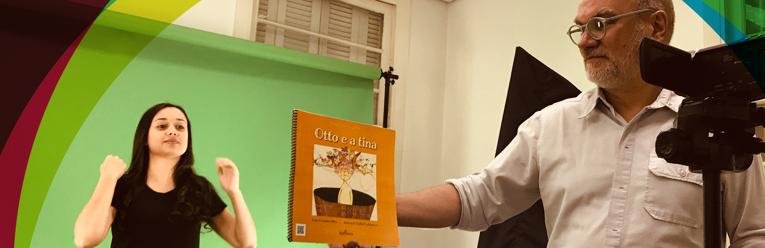 foto onde mostra um estúdio com uma mulher fazendo a língua de sinais, e um homem segurando o livro do Otto e a tina e detalhe de uma câmera fotográfica a direita da foto.